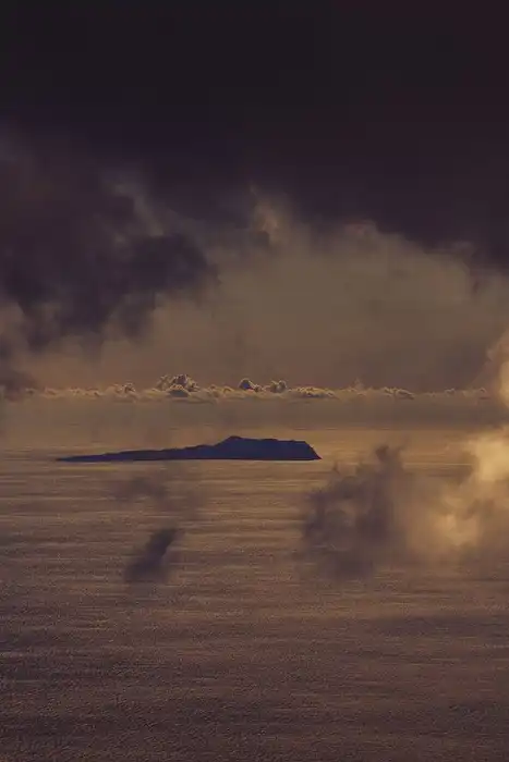 Storm clouds hang over a distant island and dark ocean beneath a band of cloud near the horizon.
