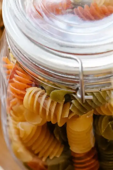 Glass jars containing dry spiral pasta and other pantry ingredients on a wooden tray.