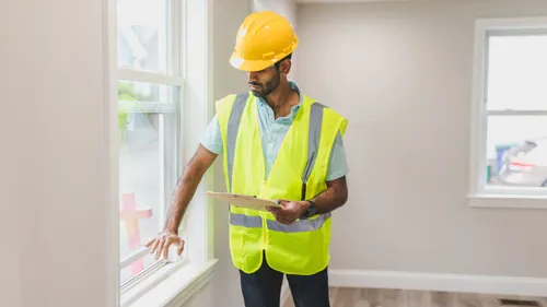 A person in a hard hat and high-visibility vest examines a residential window while holding a clipboard.