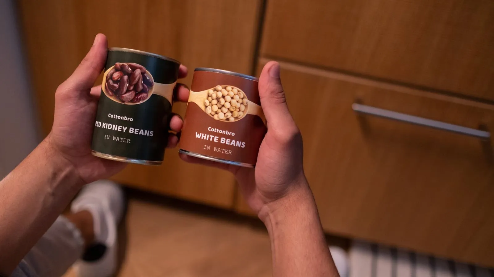 Hands holding two cans of beans in a kitchen.
