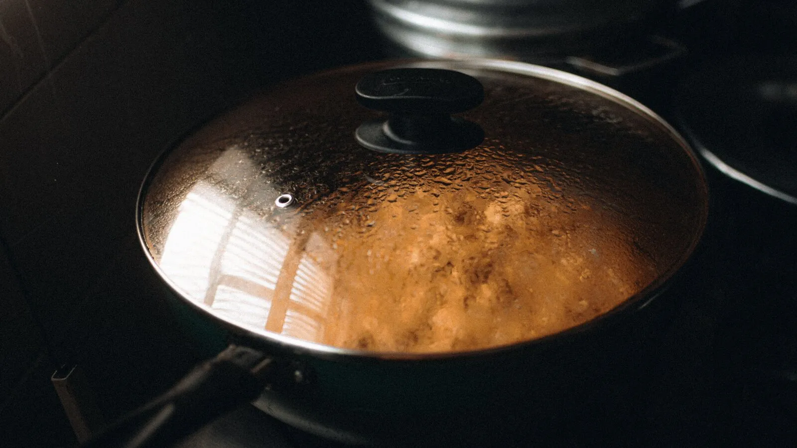 Steam beads on the glass lid of a pan sitting on a stovetop.
