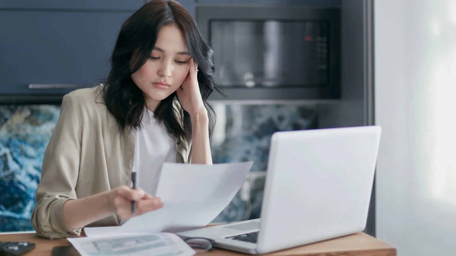 Woman reviewing a sheet of household paperwork beside a laptop at home