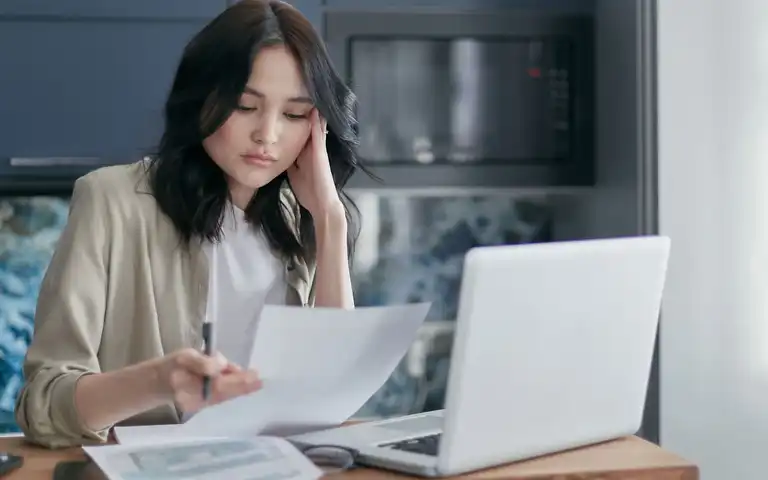 Woman reviewing a sheet of household paperwork beside a laptop at home