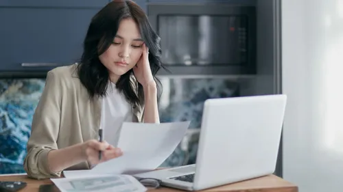 Woman reviewing a sheet of household paperwork beside a laptop at home