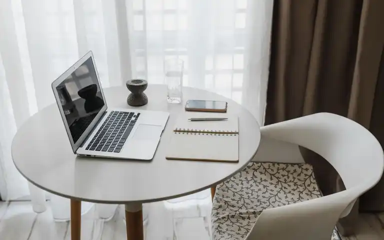 An open laptop, notebook and phone on a round table beside a bright window.