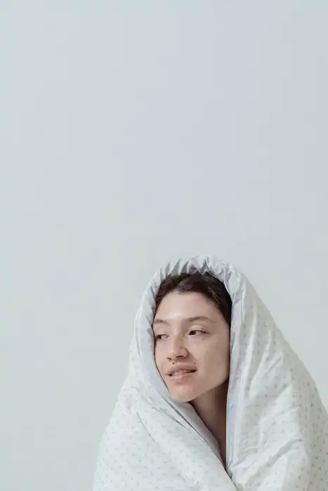 A person wrapped in a light blanket while sitting on a bed.