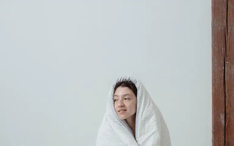 A person wrapped in a light blanket while sitting on a bed.