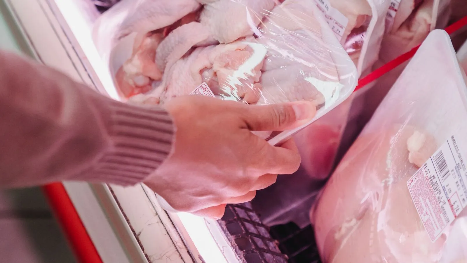 A shopper selects a tray of packaged raw chicken from a refrigerated case.
