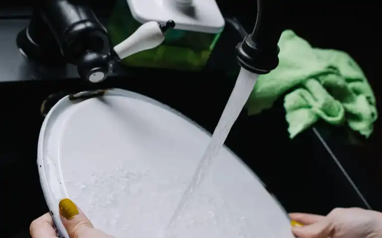 Hands wash a plate under running water in a kitchen sink.