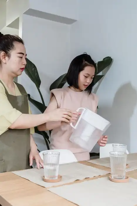 A woman and child pour water from a filter jug at a kitchen table.
