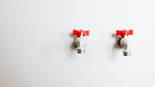 Two metal faucets with red handles are mounted on a white wall.